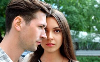 A close-up of a young man and woman standing outdoors against a backdrop of green trees. The man, with short brown hair, looks forward with a serious expression, while the woman, with long brown hair, gazes directly at the camera with a neutral expression.