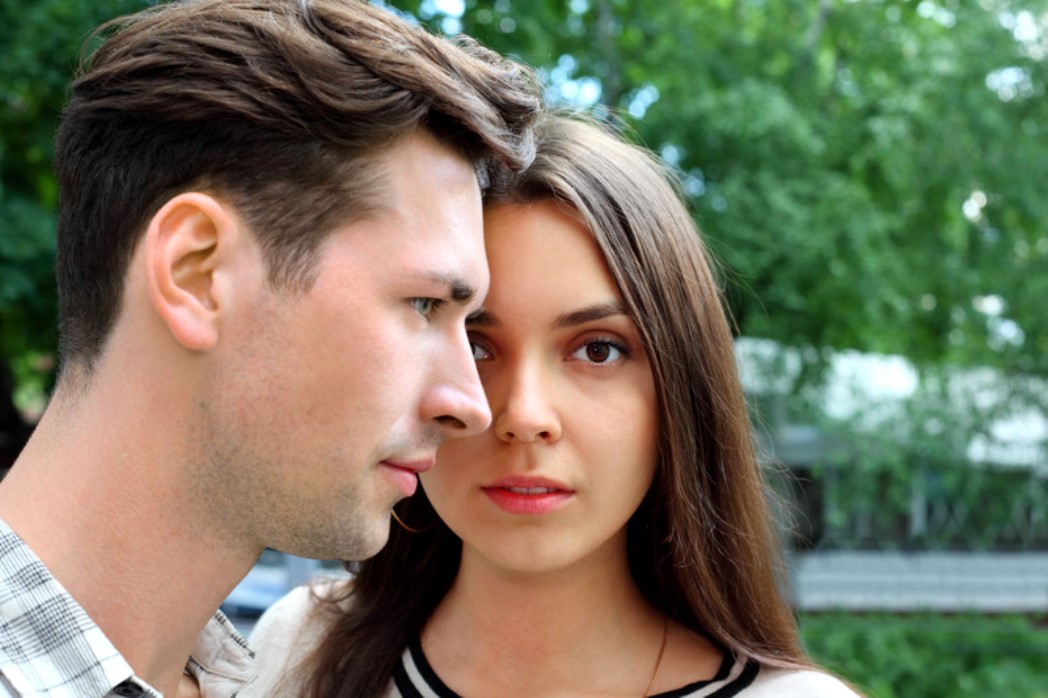 A close-up of a young man and woman standing outdoors against a backdrop of green trees. The man, with short brown hair, looks forward with a serious expression, while the woman, with long brown hair, gazes directly at the camera with a neutral expression.