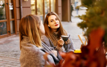 Two women sit at an outdoor café table on a cobblestone sidewalk. The woman on the right holds a cup, has long brown hair, and wears red lipstick. Both are wrapped in shawls, with autumn leaves in the foreground. The background shows buildings and a blurred street view.