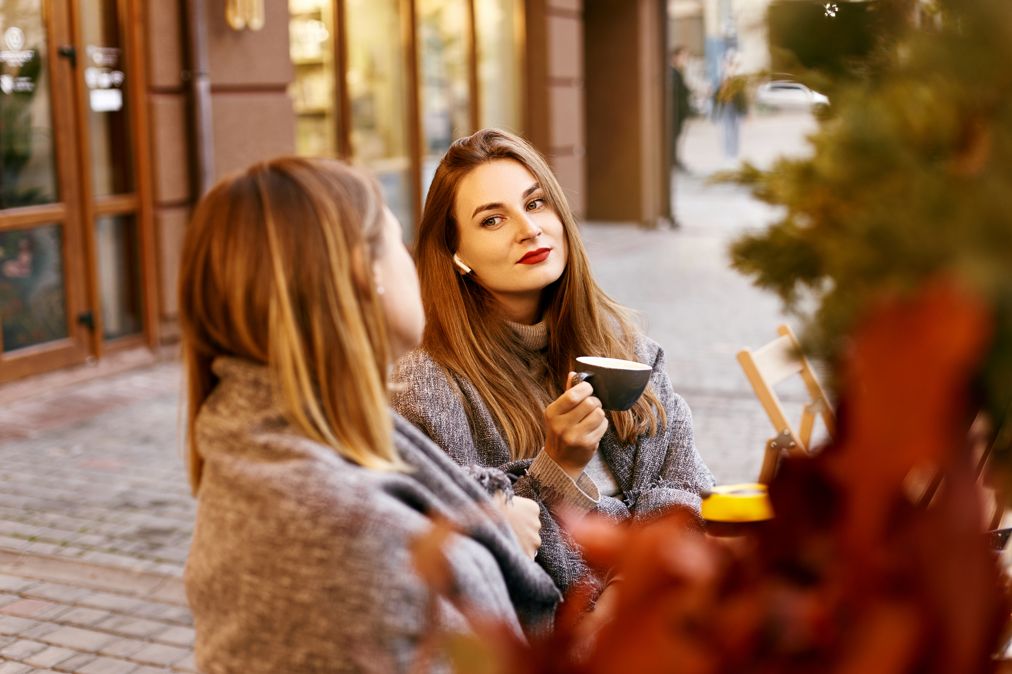 Two women sit at an outdoor café table on a cobblestone sidewalk. The woman on the right holds a cup, has long brown hair, and wears red lipstick. Both are wrapped in shawls, with autumn leaves in the foreground. The background shows buildings and a blurred street view.