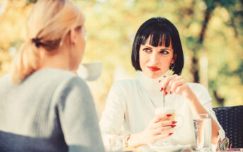 Two women are sitting outdoors at a table, engaged in conversation. One has blonde hair and is seen from the back. The other has dark hair with bangs, red lipstick, and is stirring a drink, looking attentively at the blonde woman. The background is blurred with greenery.