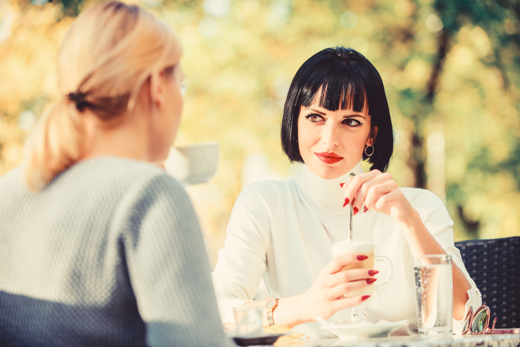 Two women are sitting outdoors at a table, engaged in conversation. One has blonde hair and is seen from the back. The other has dark hair with bangs, red lipstick, and is stirring a drink, looking attentively at the blonde woman. The background is blurred with greenery.