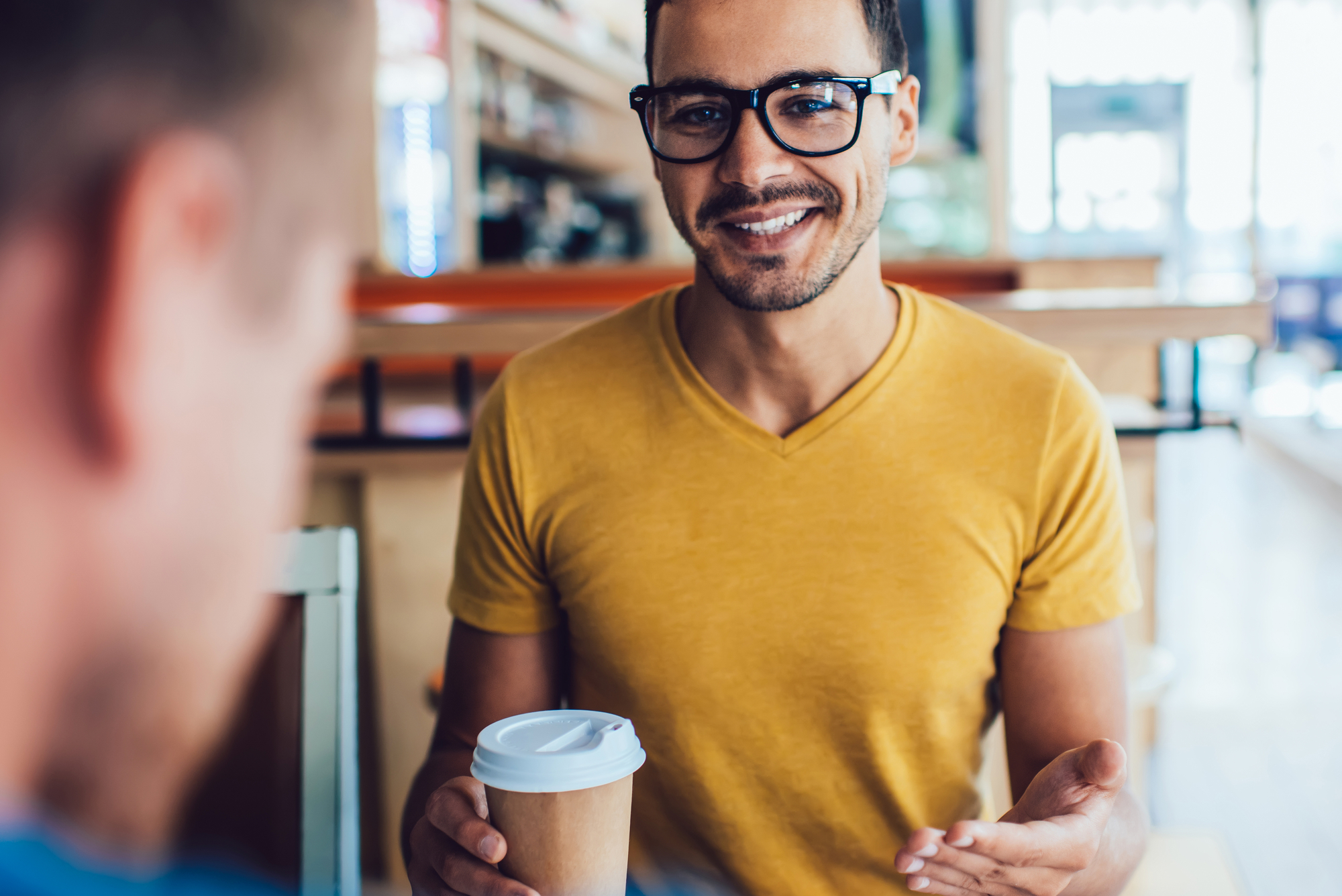 A smiling man wearing a yellow t-shirt and glasses is sitting in a café, holding a paper coffee cup. The background includes blurred café furniture and shelves. The man's body language suggests he is engaged in a conversation with someone off-camera.