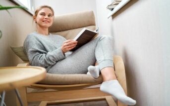 A woman wearing a grey sweatshirt and sweatpants is sitting comfortably in a cushioned wooden chair. She is holding an open book and smiling, with her legs relaxed and resting on the chair. A small round table with a plant is beside her.