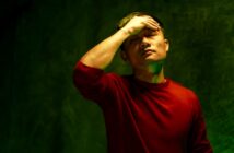 A person wearing a red shirt stands against a dark green background, with eyes closed and one hand on their forehead, appearing to be in a state of distress or deep thought.