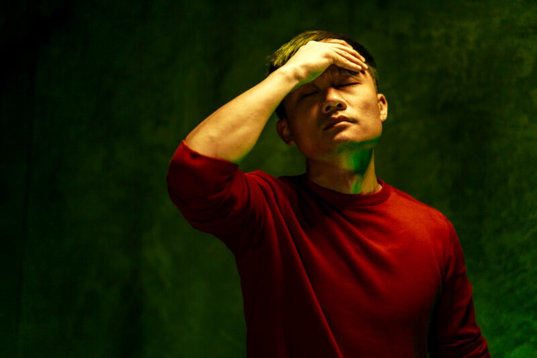 A person wearing a red shirt stands against a dark green background, with eyes closed and one hand on their forehead, appearing to be in a state of distress or deep thought.