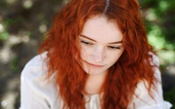 A person with long, wavy red hair and light skin looks down with a calm expression. They are wearing a white top and are outdoors, surrounded by blurred greenery. The background is softly out of focus, emphasizing the subject's serene face.