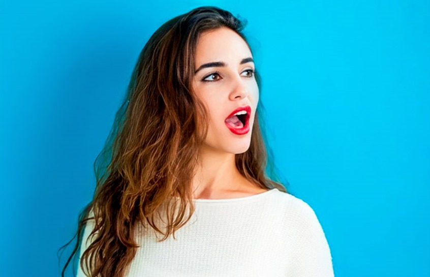 A woman with long, wavy brown hair stands against a bright blue background. She is wearing a white top and has an expression of surprise or awe, with her mouth slightly open and eyes looking off to the side.