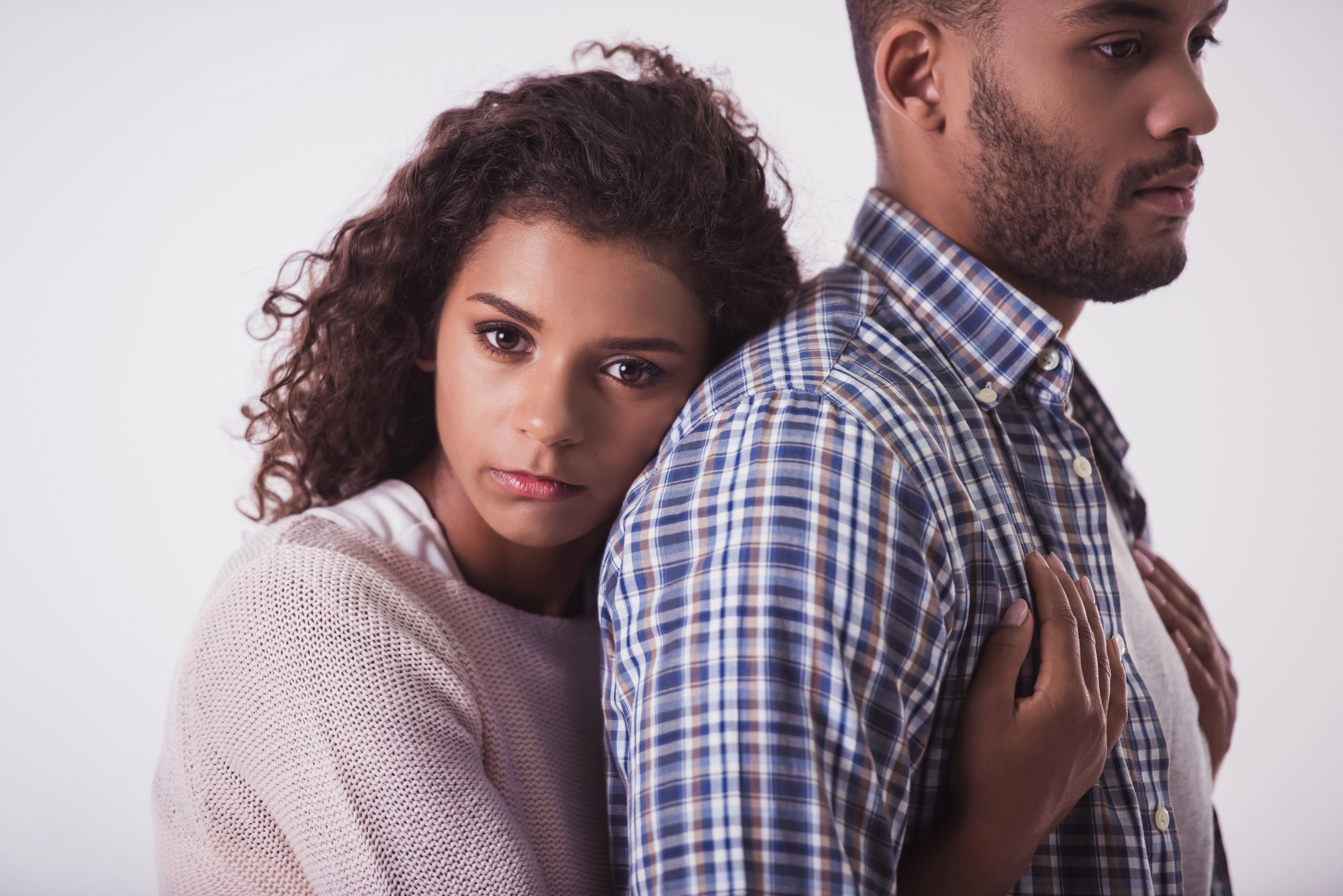 A woman with curly hair, wearing a light pink sweater, stands behind a man in a checkered shirt, resting her head on his back and holding onto his arms. The man looks forward with a serious expression. The background is plain white.