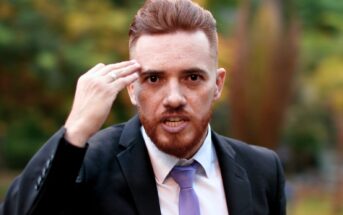 A man with a red beard and short red hair is wearing a black suit, white shirt, and lavender tie. He is outdoors, with a blurred background of greenery, gesturing with his right hand near his temple while looking slightly to the right of the camera.