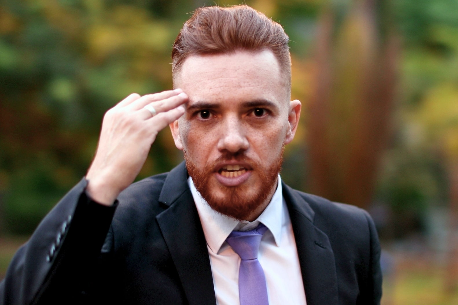 A man with a red beard and short red hair is wearing a black suit, white shirt, and lavender tie. He is outdoors, with a blurred background of greenery, gesturing with his right hand near his temple while looking slightly to the right of the camera.