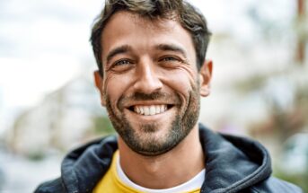 A smiling man with short dark hair and a beard is wearing a yellow shirt and a black jacket. He is standing outdoors with a blurred background of buildings and trees on a cloudy day.