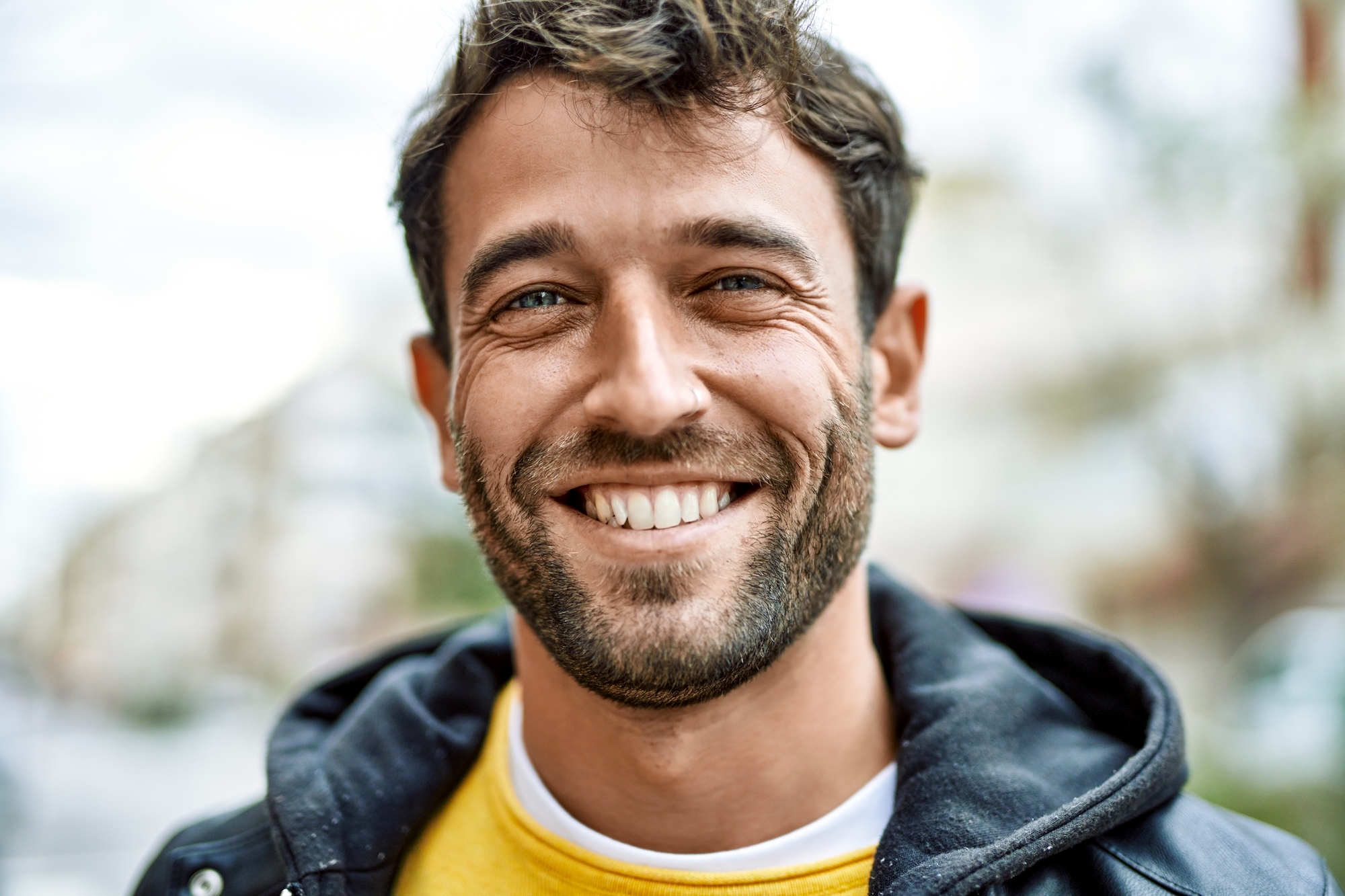 A smiling man with short dark hair and a beard is wearing a yellow shirt and a black jacket. He is standing outdoors with a blurred background of buildings and trees on a cloudy day.