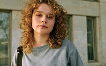 A person with curly hair and wearing a gray shirt looks directly at the camera. They have a neutral expression and are standing in front of a building with large windows. One strap of a black bag is visible over their shoulder.