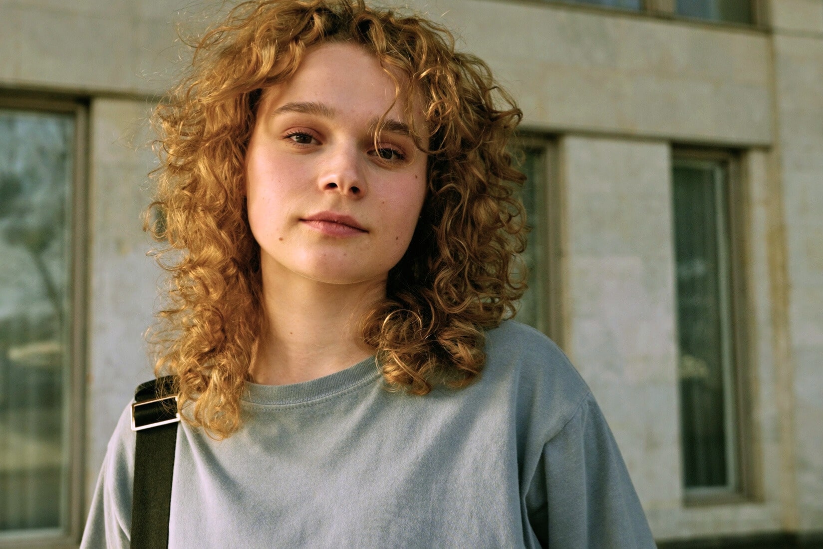 A person with curly hair and wearing a gray shirt looks directly at the camera. They have a neutral expression and are standing in front of a building with large windows. One strap of a black bag is visible over their shoulder.