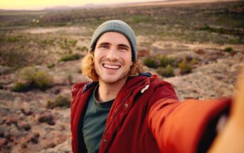 A young man with shoulder-length curly hair and wearing a green beanie and red jacket smiles at the camera while taking a selfie outdoors. The background reveals an expansive, sunlit landscape of rocky terrain and a distant horizon.