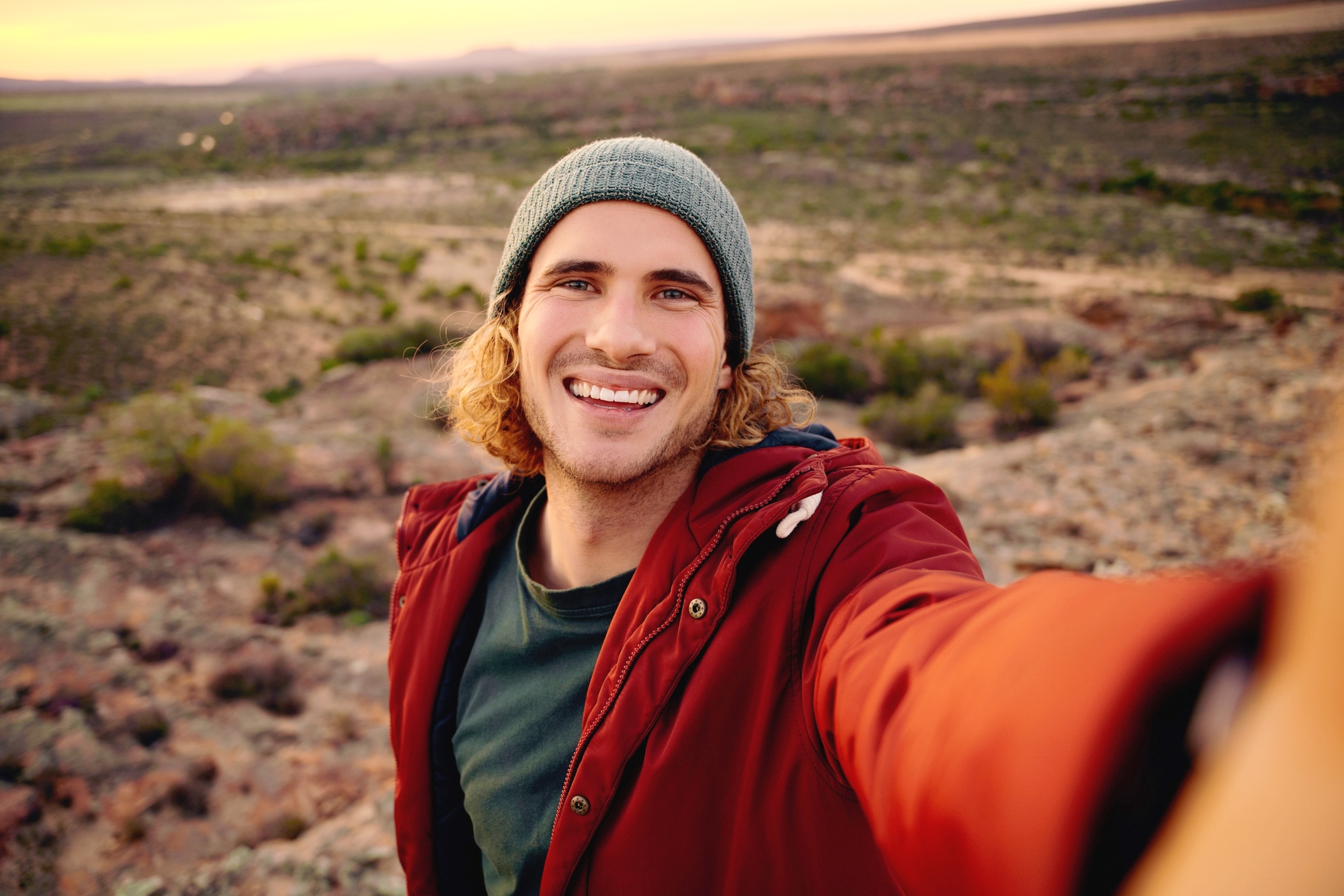 A young man with shoulder-length curly hair and wearing a green beanie and red jacket smiles at the camera while taking a selfie outdoors. The background reveals an expansive, sunlit landscape of rocky terrain and a distant horizon.