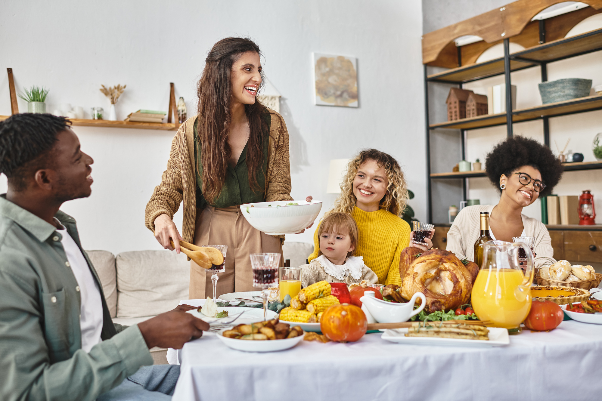 A diverse group of people sharing a Thanksgiving meal. A woman stands while serving food, smiling at the guests seated at the table, which is laden with dishes, including a roasted turkey, corn, and various side dishes. Glasses of juice and wine are also on the table.