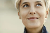 Close-up of a person with short, light-colored hair, looking slightly up and to the right with a gentle smile. The background is blurred, focusing on their face which conveys a sense of contentment and contemplation.