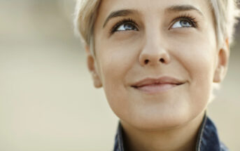 Close-up of a person with short, light-colored hair, looking slightly up and to the right with a gentle smile. The background is blurred, focusing on their face which conveys a sense of contentment and contemplation.