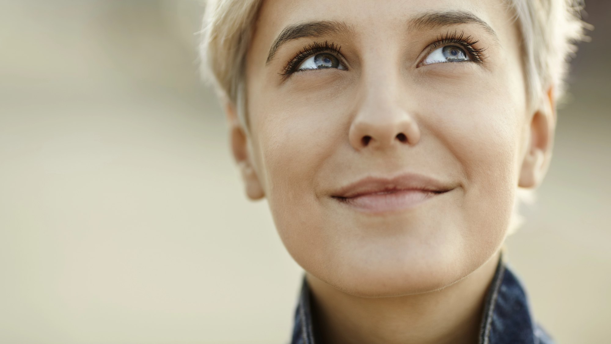 Close-up of a person with short, light-colored hair, looking slightly up and to the right with a gentle smile. The background is blurred, focusing on their face which conveys a sense of contentment and contemplation.