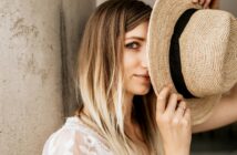 A woman with long, blonde hair partially covers her face with a straw hat. She looks towards the camera with one eye visible. She is dressed in a white lace top and stands by a concrete column. Natural light illuminates the scene.