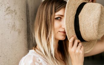 A woman with long, blonde hair partially covers her face with a straw hat. She looks towards the camera with one eye visible. She is dressed in a white lace top and stands by a concrete column. Natural light illuminates the scene.