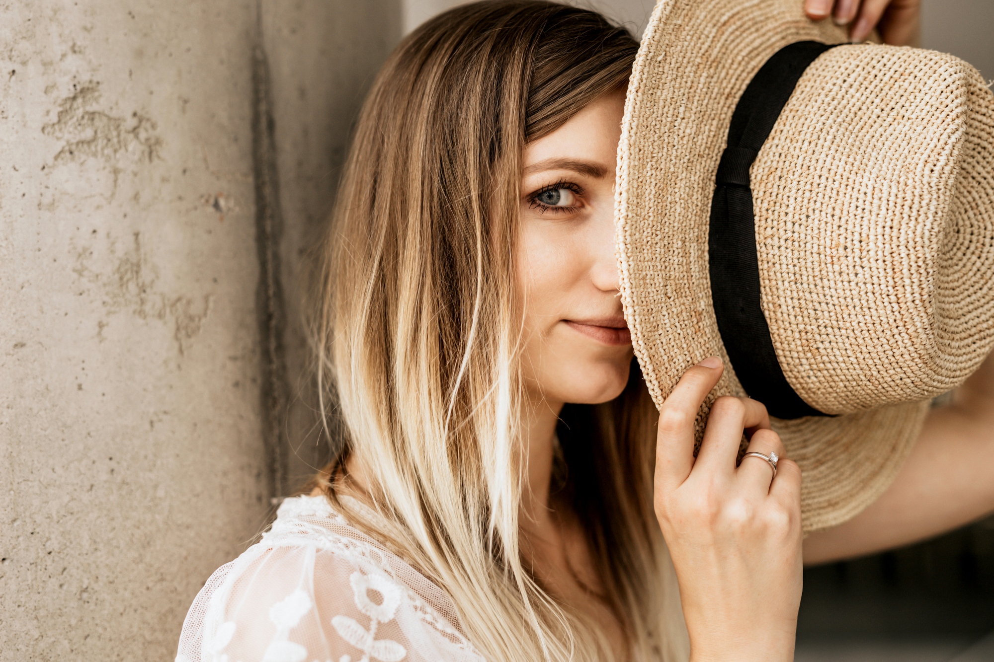 A woman with long, blonde hair partially covers her face with a straw hat. She looks towards the camera with one eye visible. She is dressed in a white lace top and stands by a concrete column. Natural light illuminates the scene.