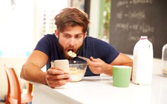 A man with a beard is eating a bowl of cereal while looking at his smartphone. He is sitting in a kitchen with a bottle of milk and a green mug on the counter. A blackboard with writing in the background is partially visible.