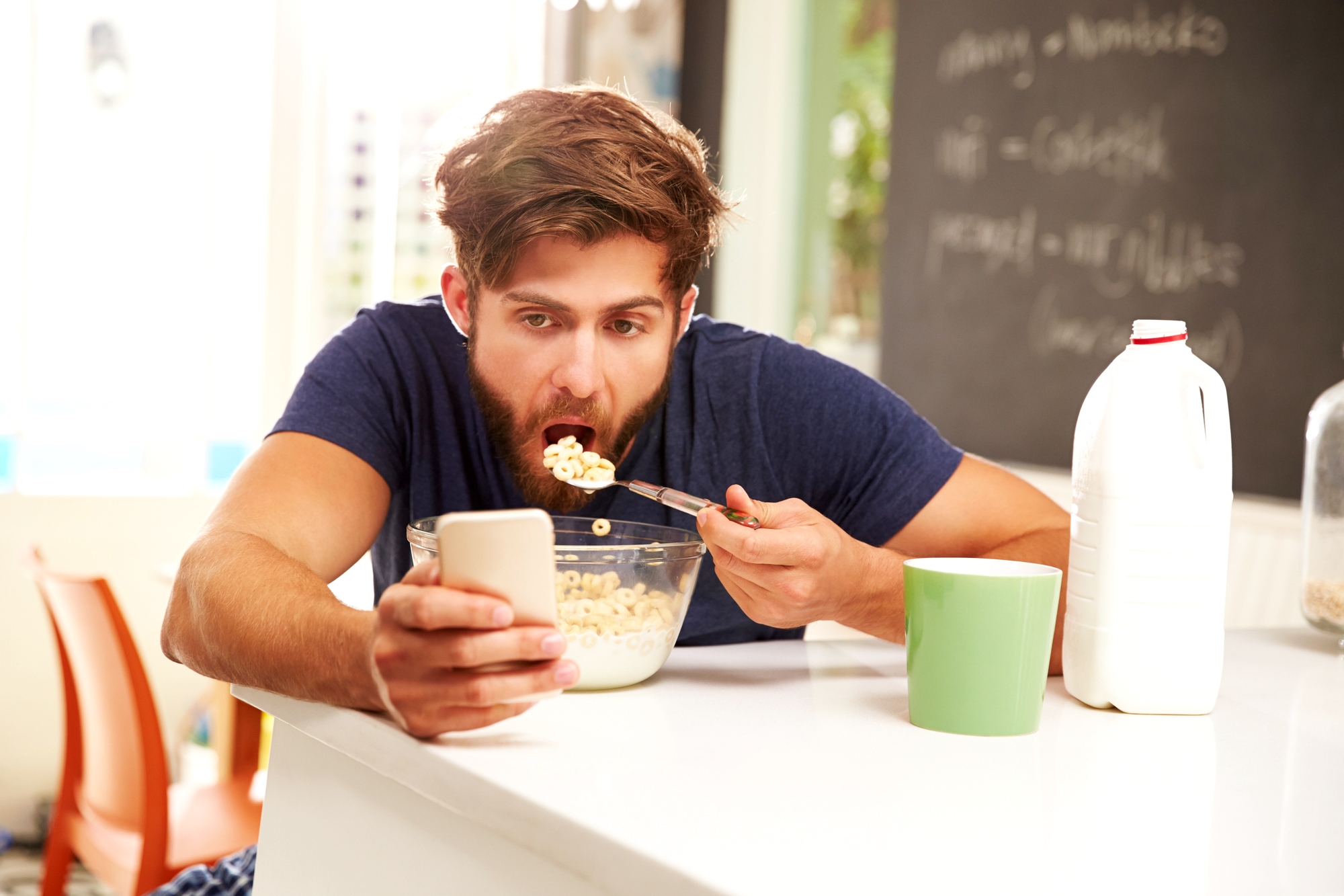 A man with a beard is eating a bowl of cereal while looking at his smartphone. He is sitting in a kitchen with a bottle of milk and a green mug on the counter. A blackboard with writing in the background is partially visible.