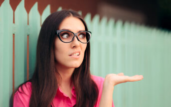 A woman with long dark hair wearing glasses and a pink shirt stands in front of a turquoise fence. Her facial expression shows confusion or bewilderment, and she has her left hand raised with the palm up as if gesturing or questioning.