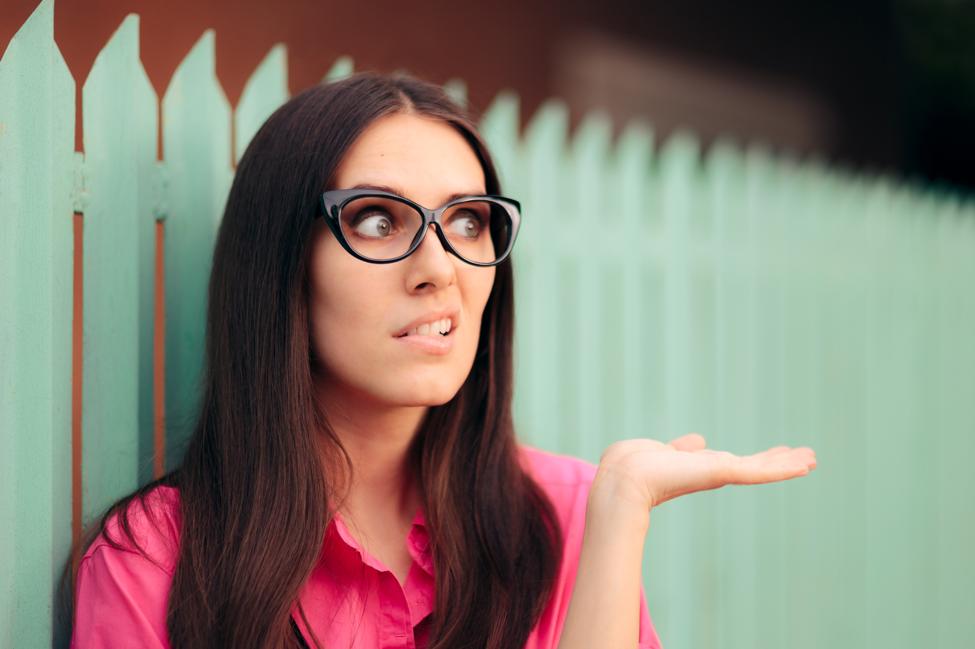 A woman with long dark hair wearing glasses and a pink shirt stands in front of a turquoise fence. Her facial expression shows confusion or bewilderment, and she has her left hand raised with the palm up as if gesturing or questioning.