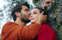 A man in an orange sweater gently kisses the forehead of a woman in a red sweater. The woman leans against a tree while looking into the distance, with the man holding her head tenderly. The background has blurred natural elements, suggesting an outdoor setting.