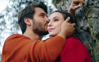 A man in an orange sweater gently kisses the forehead of a woman in a red sweater. The woman leans against a tree while looking into the distance, with the man holding her head tenderly. The background has blurred natural elements, suggesting an outdoor setting.