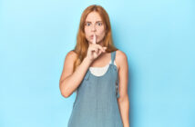 A woman with long red hair stands against a light blue background, wearing a sleeveless blue top. She is holding a finger to her lips in a "shhhh" gesture, looking directly at the camera with wide eyes.