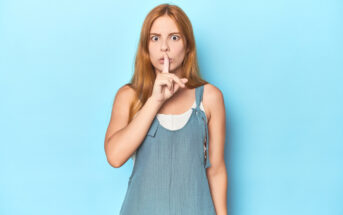 A woman with long red hair stands against a light blue background, wearing a sleeveless blue top. She is holding a finger to her lips in a "shhhh" gesture, looking directly at the camera with wide eyes.