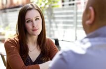 A woman with long brown hair and wearing a brown cardigan is seated outdoors, looking attentively towards a bald man in a blue shirt whose back is to the camera. They appear to be engaged in a conversation. A blurred fence and greenery are visible in the background.