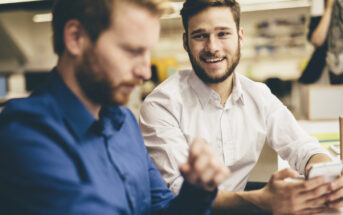 Two men are sitting in an office. The man on the left, wearing a blue shirt, is looking down while the man on the right, in a white shirt, is smiling, holding a smartphone. The background is blurred, with light coming from overhead lamps and a computer screen visible.