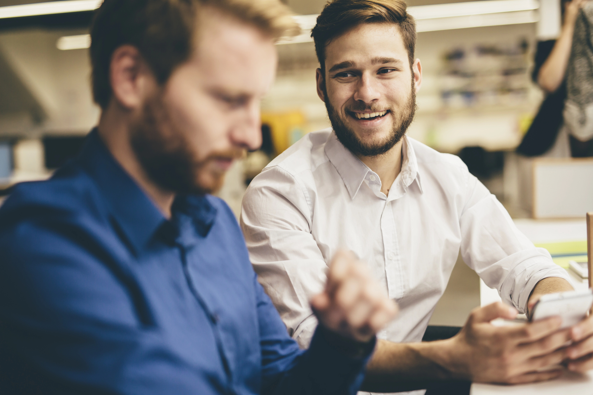 Two men are sitting in an office. The man on the left, wearing a blue shirt, is looking down while the man on the right, in a white shirt, is smiling, holding a smartphone. The background is blurred, with light coming from overhead lamps and a computer screen visible.