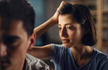 A woman with short brown hair and a sad expression places her hand on the shoulder of a man, who is facing away from her. The man appears distant, and their body language suggests tension or emotional discomfort. The setting is indoors with a blurred background.