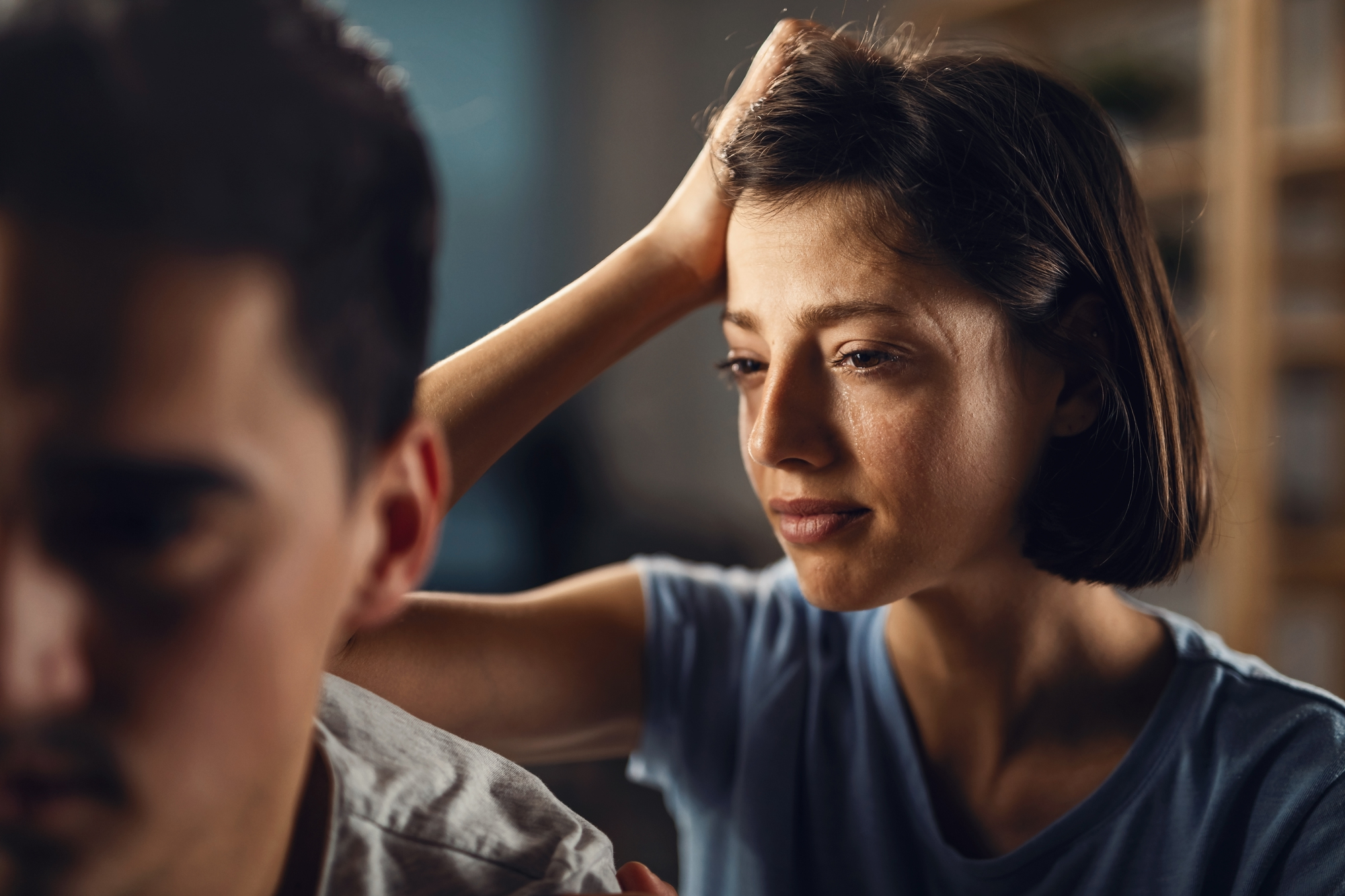 A woman with short brown hair and a sad expression places her hand on the shoulder of a man, who is facing away from her. The man appears distant, and their body language suggests tension or emotional discomfort. The setting is indoors with a blurred background.