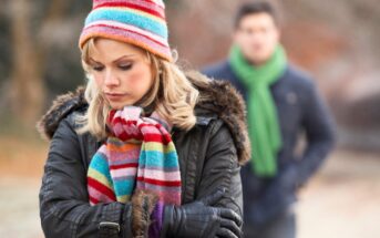 A woman wearing a colorful striped hat and scarf stands with her arms crossed and a sad expression. A man in the background, wearing a green scarf and dark jacket, appears out of focus. The scene is outdoors in a cold setting.