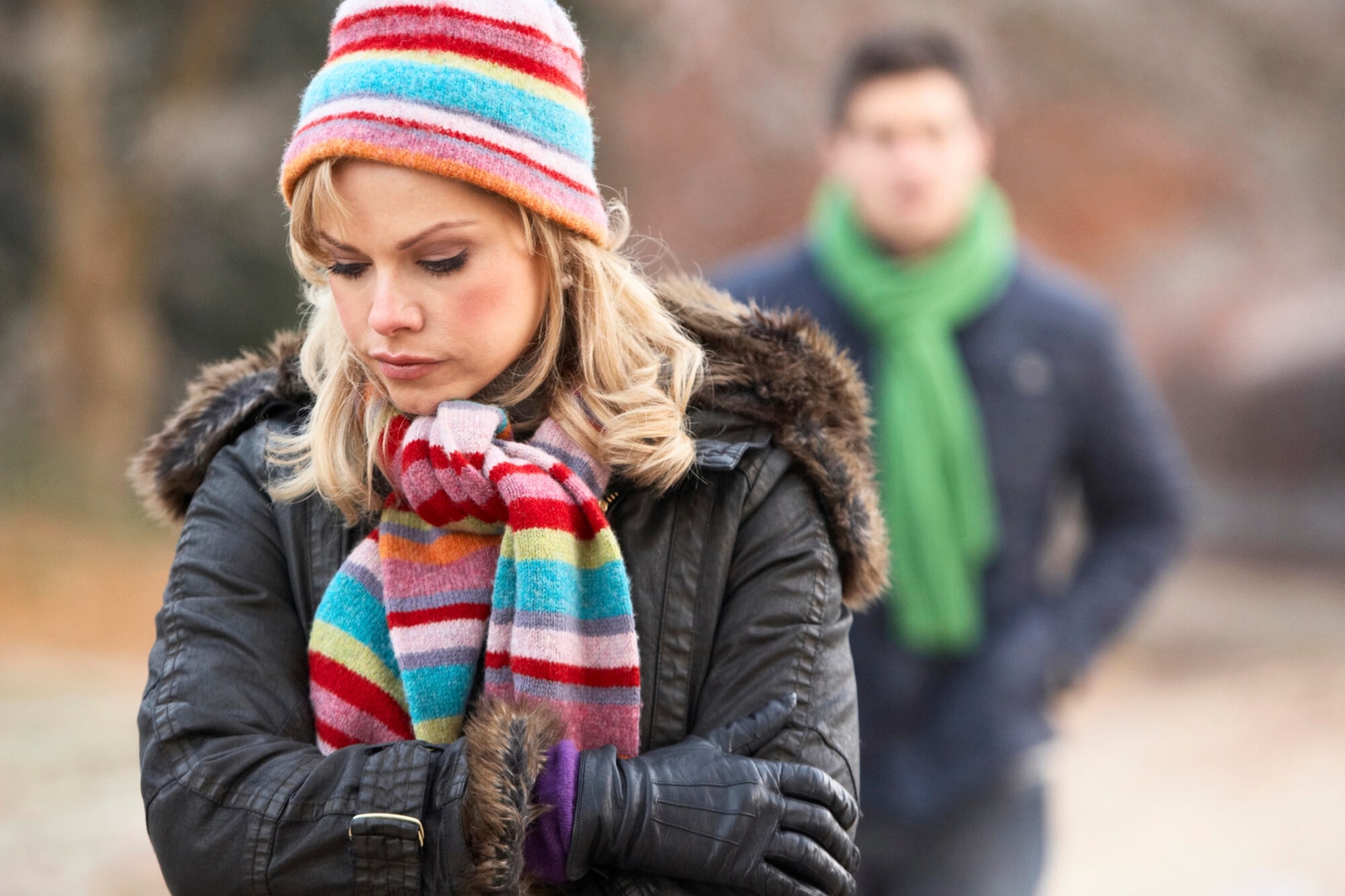 A woman wearing a colorful striped hat and scarf stands with her arms crossed and a sad expression. A man in the background, wearing a green scarf and dark jacket, appears out of focus. The scene is outdoors in a cold setting.