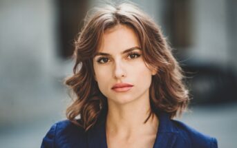 A woman with wavy brown hair and a serious expression is looking directly at the camera. She is wearing a dark-colored blazer. The background is blurred, highlighting the focus on her face.