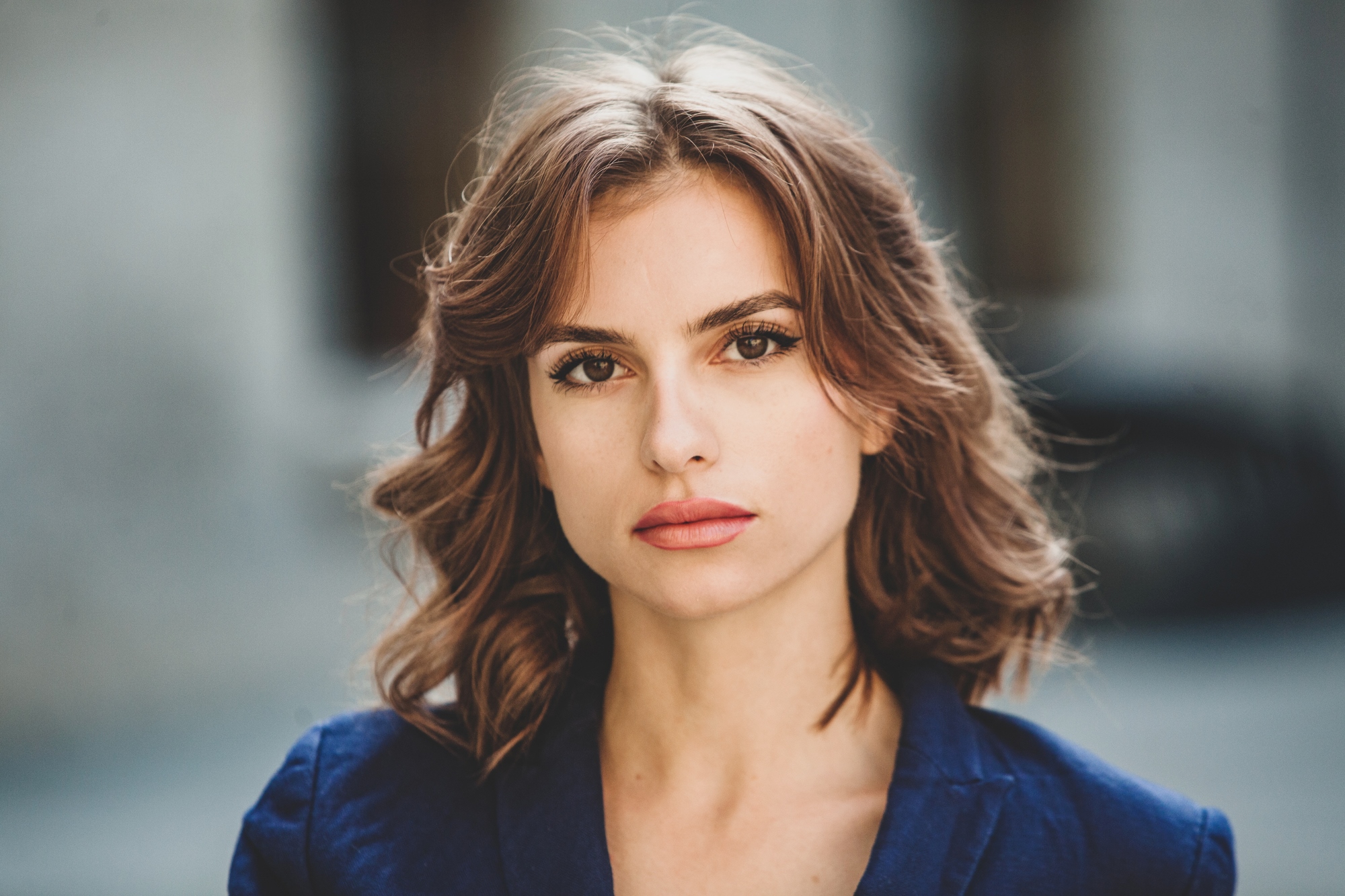 A woman with wavy brown hair and a serious expression is looking directly at the camera. She is wearing a dark-colored blazer. The background is blurred, highlighting the focus on her face.