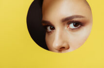 A close-up of a woman's eyes and eyebrows peeking through a circular hole in a yellow background. The lighting highlights her well-groomed eyebrows and long lashes, creating a striking contrast against the bright yellow.