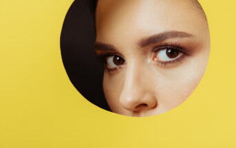 A close-up of a woman's eyes and eyebrows peeking through a circular hole in a yellow background. The lighting highlights her well-groomed eyebrows and long lashes, creating a striking contrast against the bright yellow.