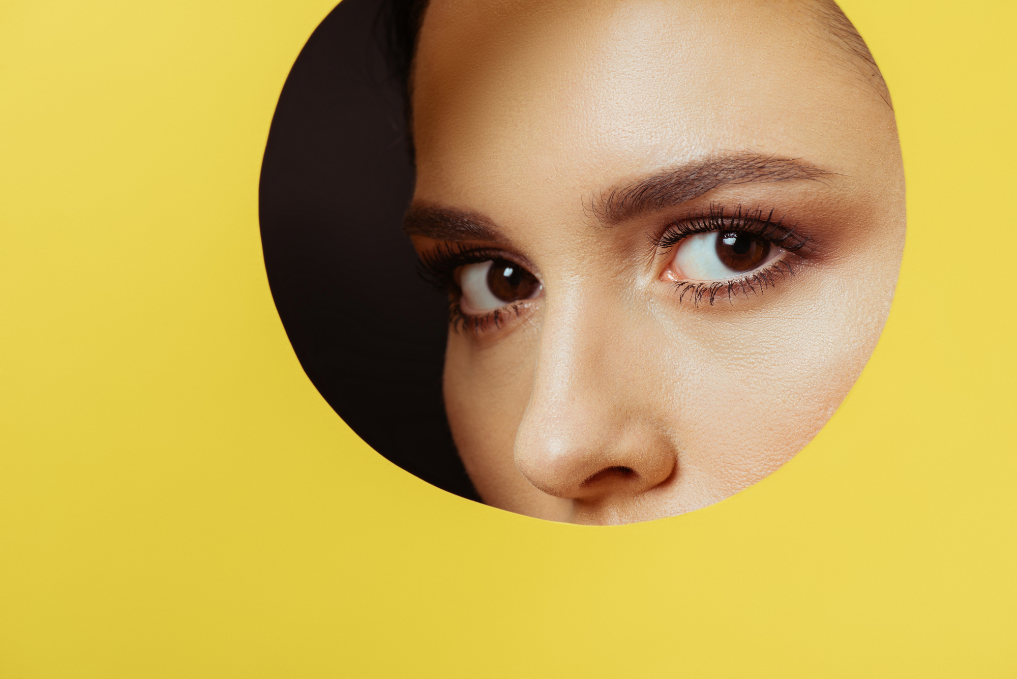 A close-up of a woman's eyes and eyebrows peeking through a circular hole in a yellow background. The lighting highlights her well-groomed eyebrows and long lashes, creating a striking contrast against the bright yellow.