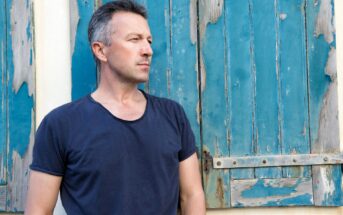 A man with short gray hair, wearing a dark blue t-shirt, stands in front of a weathered wooden wall with peeling blue paint. He is looking to the side, appearing contemplative.