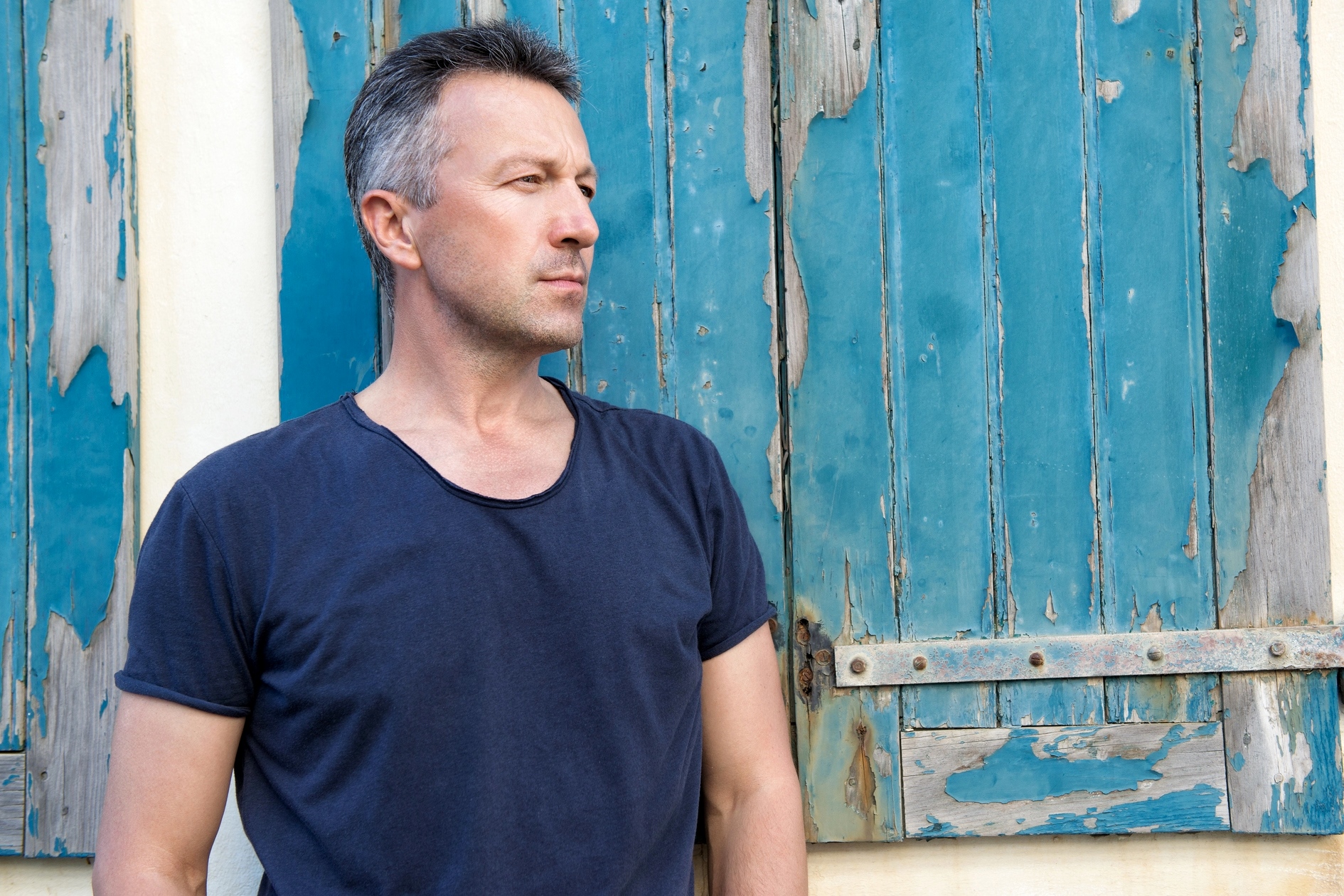 A man with short gray hair, wearing a dark blue t-shirt, stands in front of a weathered wooden wall with peeling blue paint. He is looking to the side, appearing contemplative.